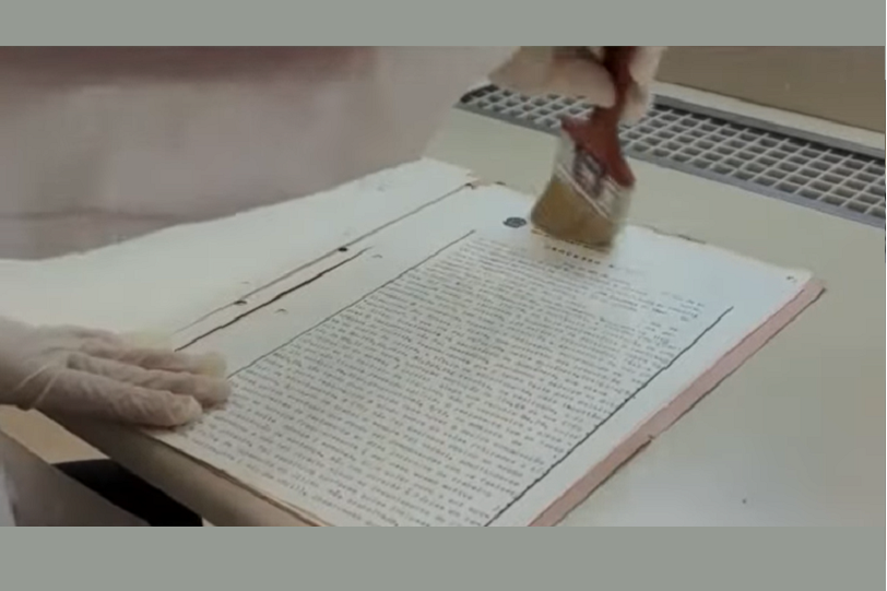 Captura de tela mostra mãos de um servidor ou servidora, de luvas brancas, segurando um pincel. Ele ou ela está limpando a página de um processo antigo.