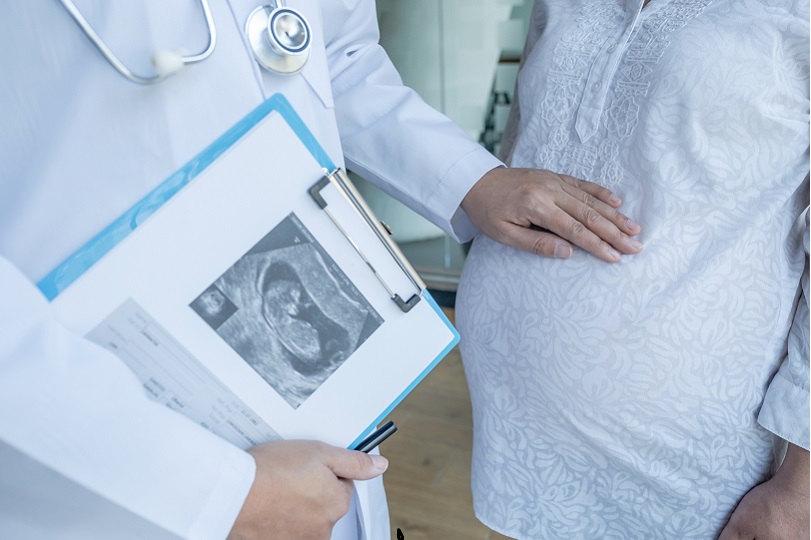 Foto ilustrativa mostra mão de médico sobre a barriga de uma grávida. Ele segura exames que mostram a imagem do bebê. Ele usa jaleco branco e estetoscópio. Ela veste camisa branca.