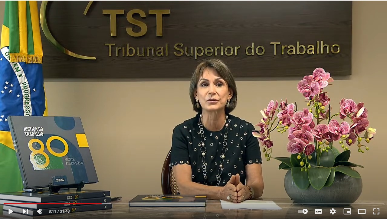 Ministra Maria Cristina Peduzzi, presidente do TST, sentada diante se uma mesa, com a logo do TST atrás e um livro dos 80 anos da Justiça do Trabalho sob a mesa. Foto: TST