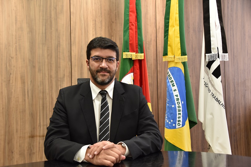 Desembargador presidente do TRT4 Francisco Rossal de Araújo sentado e com antebraços apoiados em uma mesa, em pose formal  junto às bandeiras do RS, Brasil e do TRT4 ao fundo.