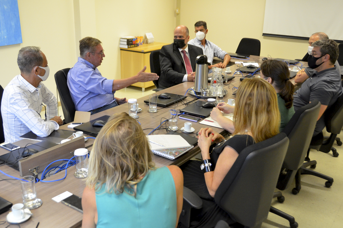 Foto com os participantes na mesa de reunião. O secretário está falando e gesticulando enquanto os outros escutam.