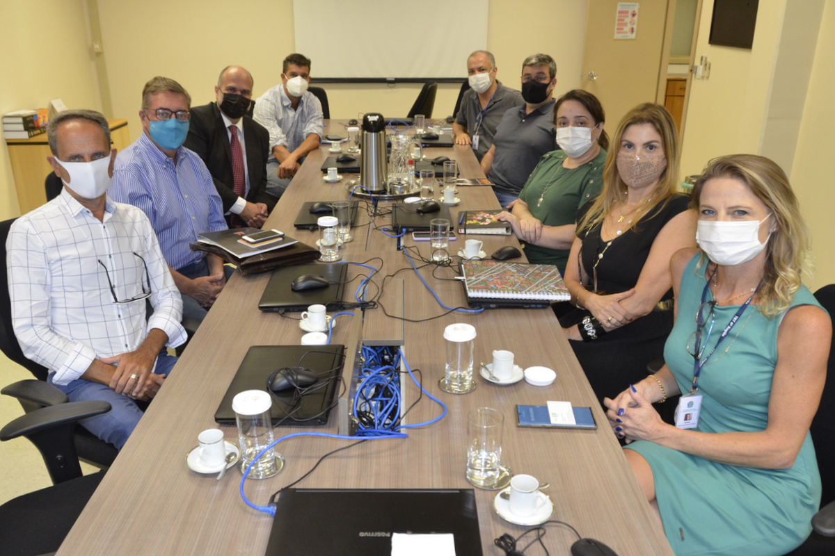 Participantes da reunião estão sentados em torno da mesa olhando para a câmera. Todos usam máscara.