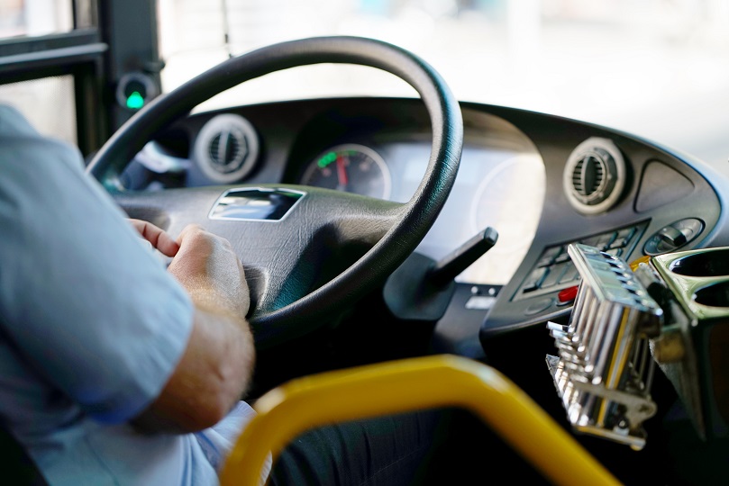Foto ilustrativa mostra mãos de motorista de ônibus sobre o volante e painel do veículo. O motorista veste camisa azul clara e a cor do painel é predominantemente preta.
