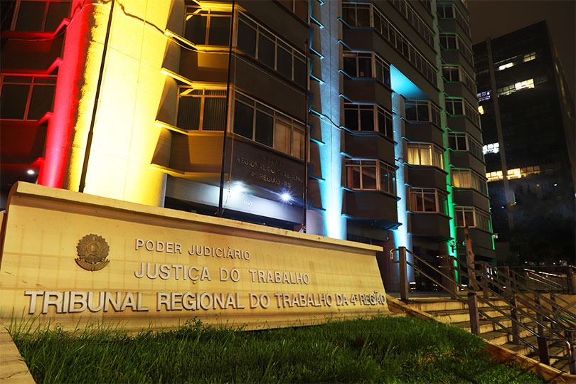 Fachada do Prédio-Sede com a iluminação oficial. Em primeiro plano aparece o letreiro com identificação do TRT-4 e ao fundo o prédio iluminado com listras verticais em vermelho, amarelo, azul e verde.