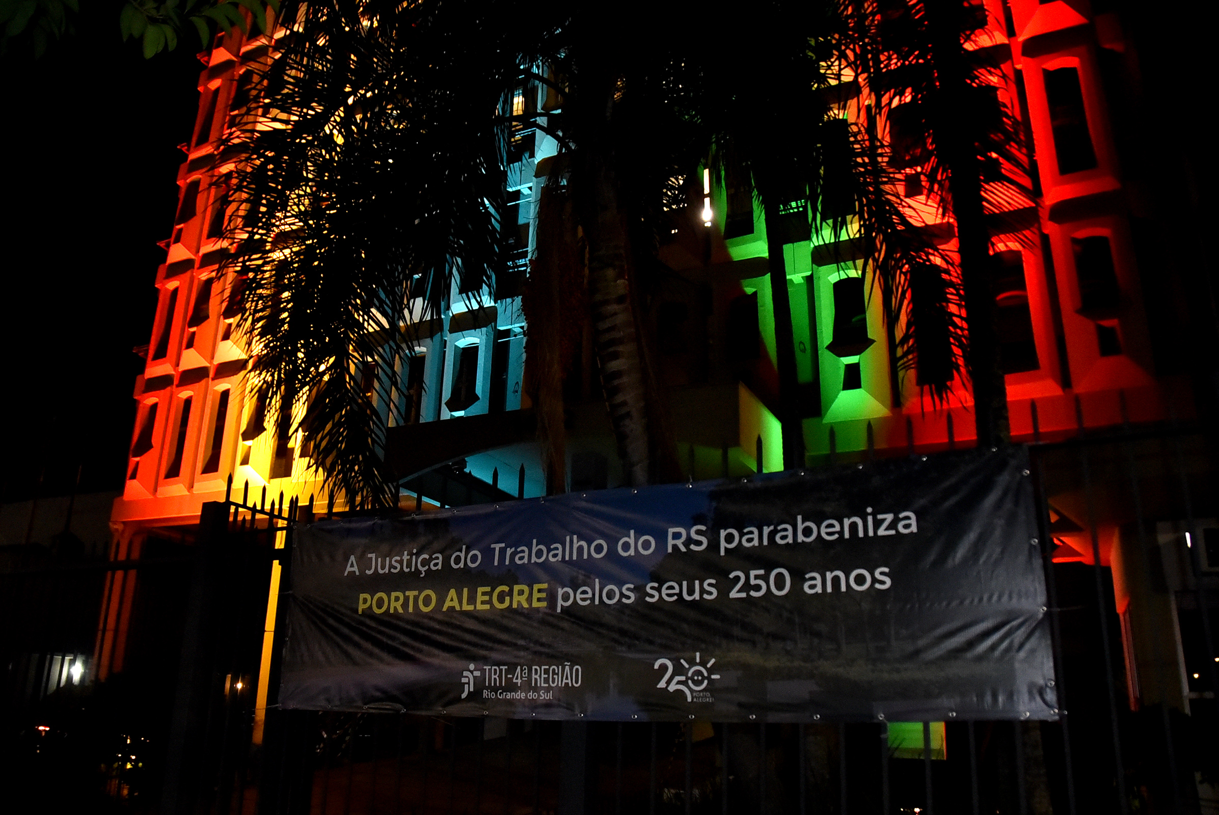 Fachada do Foro Trabalhista com a iluminação especial. Na grade em frente ao prédio aparece uma faixa com o texto "A Justiça do Trabalho gaúcha parabeniza Porto Alegre pelos seus 250 anos"