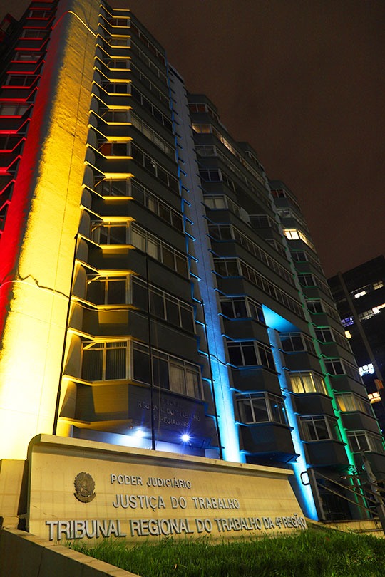 Fachada do Prédio-Sede com a iluminação oficial. Em primeiro plano aparece o letreiro e ao fundo o prédio iluminado. Imagem de baixo para cima mostra toda a altura do prédio.