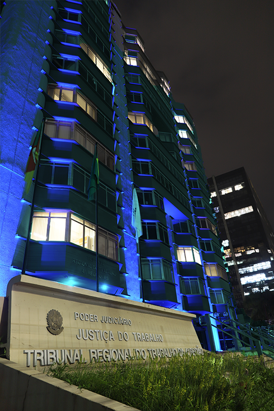 Fachada do prédio-sede do TRT-RS, iluminada na cor azul. A foto é de baixo para cima, mostra o letreiro da Justiça do Trabalho e as janelas da frente do edifício.