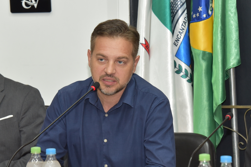 Jonas Calvi é branco, usa cabelo e barba curta e uma camisa social azul. 