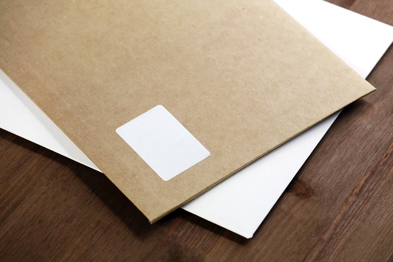 Foto ilustrativa mostra um envelope de cor parda sobre outro branco. Ambos estão sobre uma mesa de madeira.