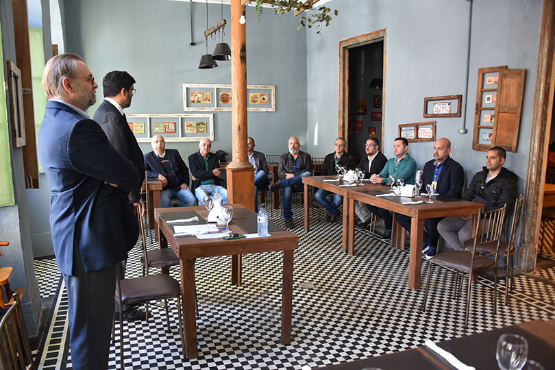 Foto em ângulo aberto da reunião, mostrando os magistrados em pé, em primeiro plano, e as demais pessoas sentadas à mesa, ao fundo.