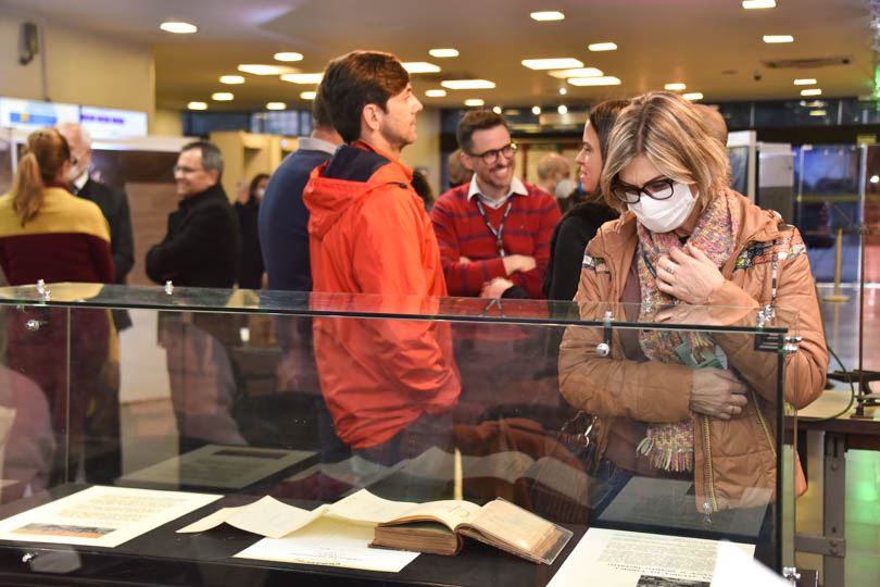 Foto do público na exposição. Uma mulher observa um livro exposto e um grupo de pessoas conversam ao fundo. 