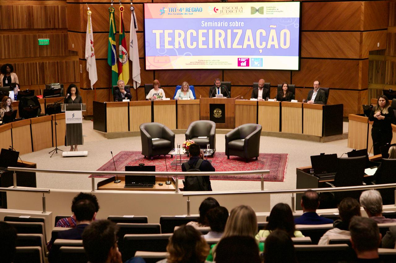 Foto em ângulo aberto do evento, mostrando público e mesa do plenário