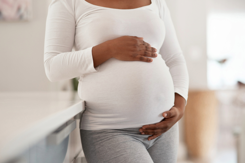 Gestante com as mãos na barriga. Ela tem a pele negra e usa blusa branca. Ao fundo, há um quarto de bebê.