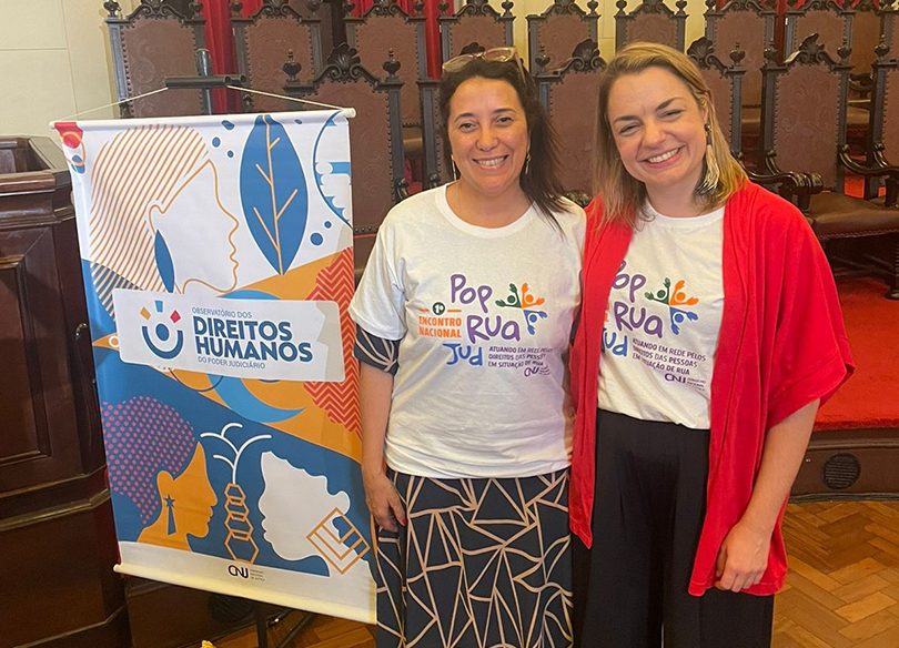 Juízas Maria Teresa e Gabriela Lacerda posam para a foto ao lado de um cartaz sobre Direitos Humanos. Elas usam camisetas com o logo do encontro.