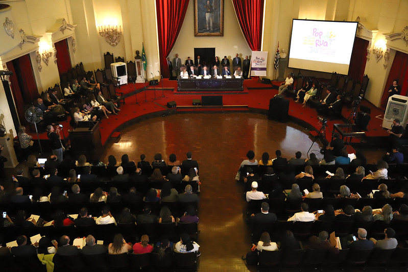 Foto geral do evento, de cima para baixo, com a mesa oficial ao fundo e plateia na parte de baixo da imagem.