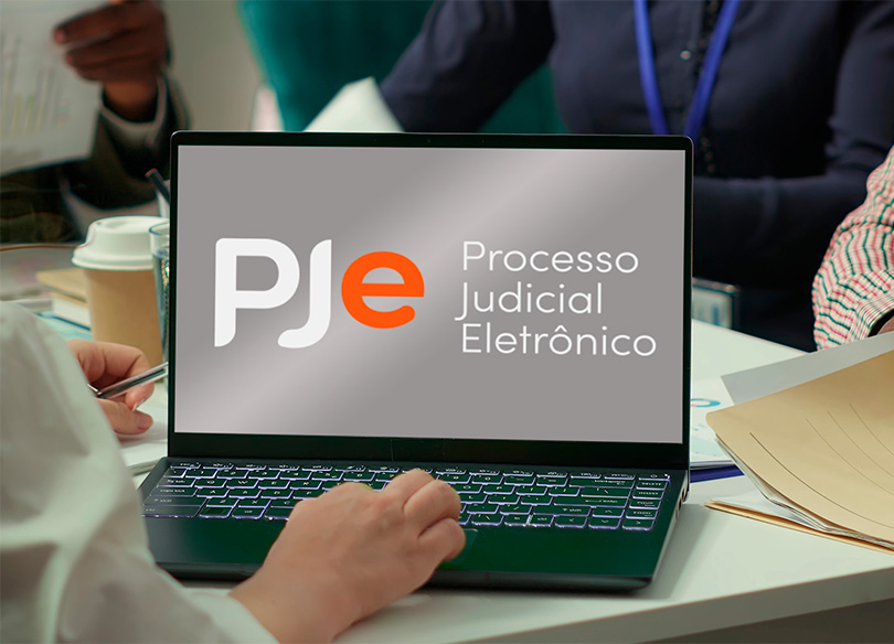 Foto de uma equipe reunida, com um notebook aberto sobre a mesa. Na tela aprece o logo do PJe.