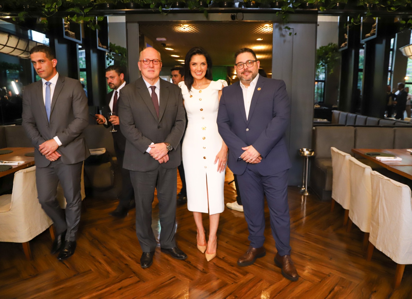 Na imagem, estão lado a lado três pessoas em um ambiente interno elegante, com piso de madeira e decoração moderna ao fundo. À esquerda, Camilo Macedo veste terno cinza e gravata azul. No centro, Márcia Helena Somensi usa um vestido branco com detalhes dourados e sorri suavemente. À direita, o desembargador Ricardo Martins Costa está de terno escuro e óculos. Todos posam de forma formal.