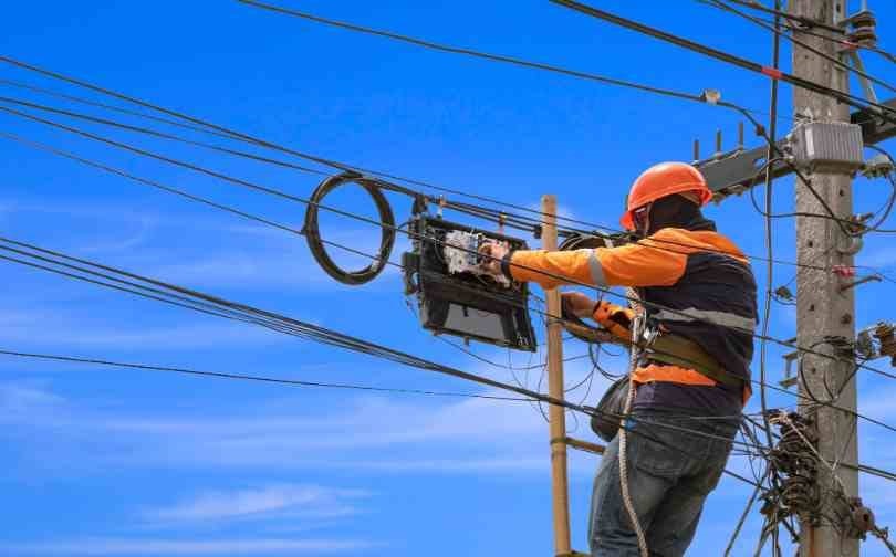 Homem faz instalação em poste. Ele veste colete preto e camisa e capacete laranja