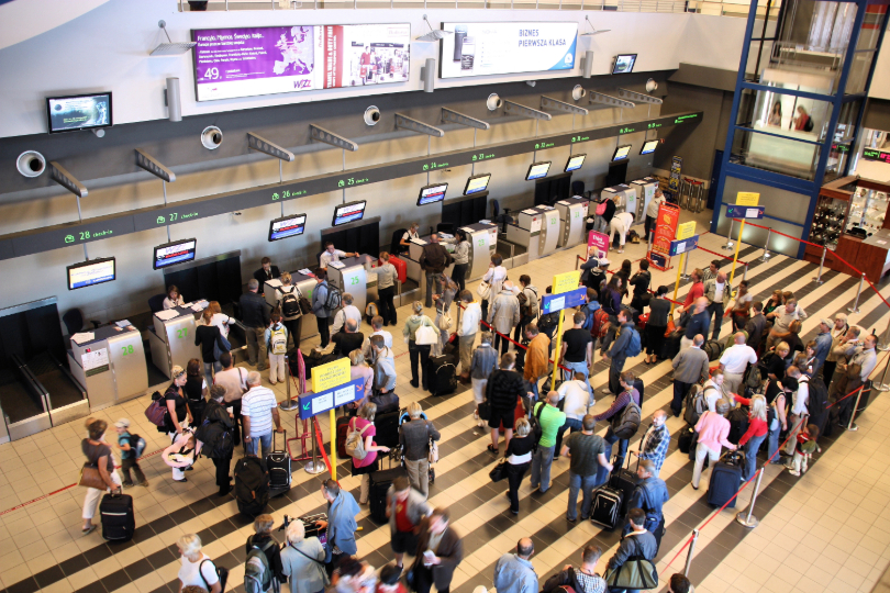Saguão de aeroporto lotado. Passageiros estão em filas para despachar bagagens.