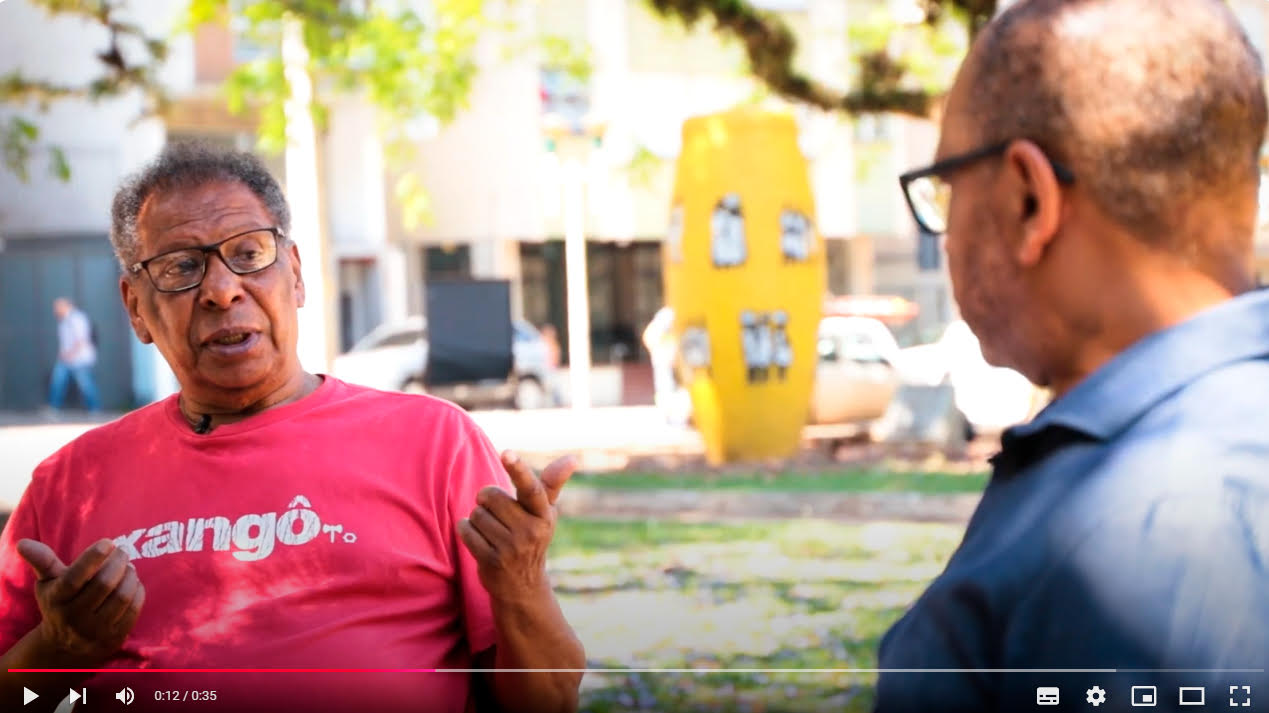 Frame do vídeo com Antônio Côrtes falando para um homem de costas, que está fazendo a entrevista. Ambos são negros. Côrtes está gesticulando. Ele usa óculos de aro preto e camiseta vermelha com a palavra "Xangô" em letras brancas. Ao fundo aparece a paisagem da praça onde eles estão, com uma escultura que representa um tambor amarelo a centro.