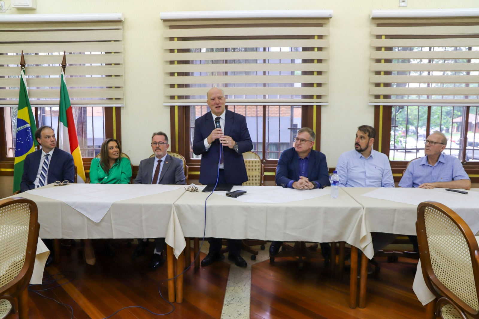 Imagem de Martins Costa com um microfone em mãos, palestrando. Ele está entre algumas pessoas num mesa oficial do evento. O evento é da CDL de Santo Ângelo.