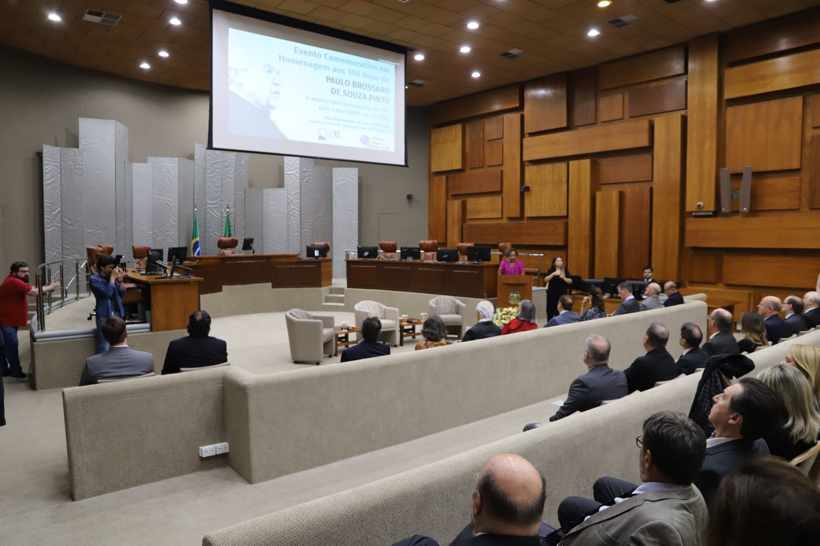A imagem captura uma plateia atenta no auditório do TRE. Em primeiro plano, três palestrantes sentados em poltronas participam de uma conversa, enquanto o público, composto por homens e mulheres em trajes formais, ocupa várias fileiras de cadeiras. A cena é de um painel em andamento.