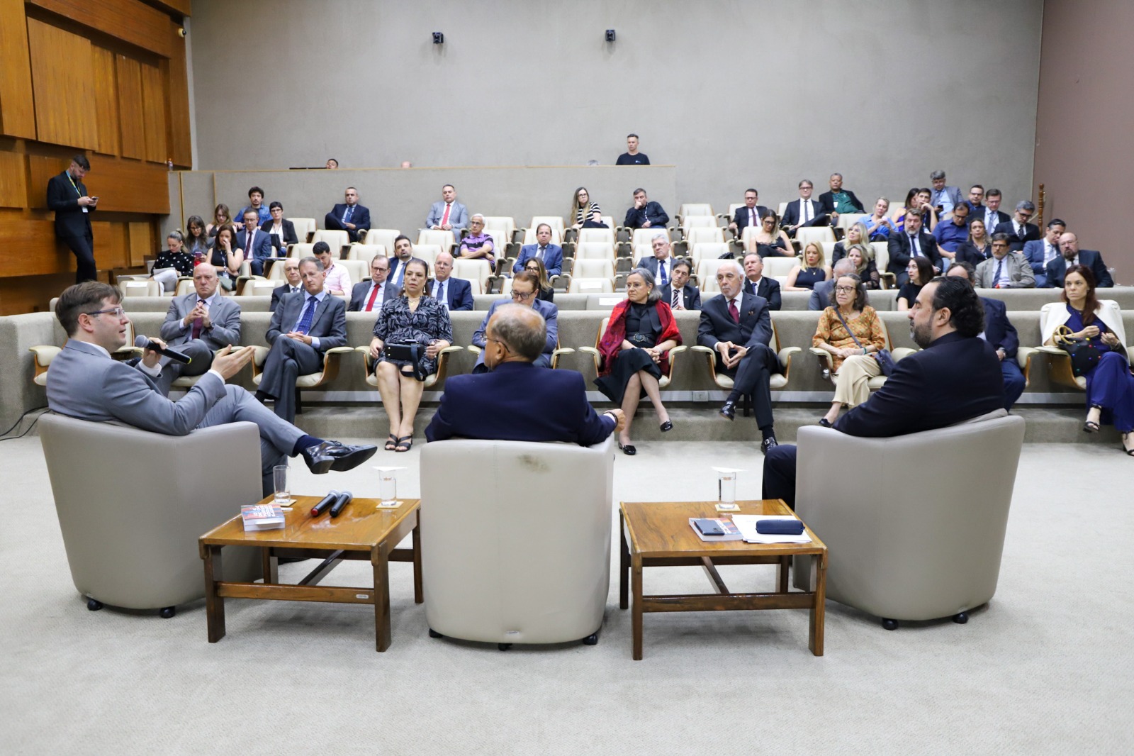 A imagem mostra o auditório do TRE, com painéis de madeira e estrutura formal. No centro, uma tela exibe informações do evento comemorativo em homenagem aos 100 anos de Paulo Brossard de Souza Pinto. Algumas pessoas estão de pé fotografando ou filmando, enquanto outras assistem sentadas. No canto direito, uma pessoa faz uso de linguagem de sinais.