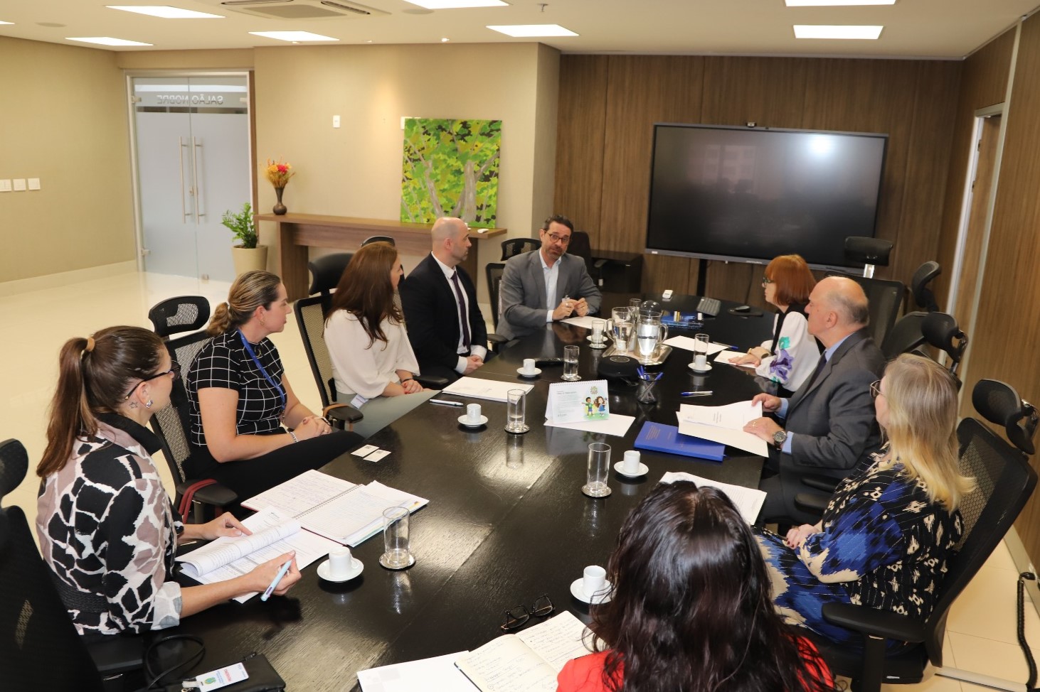 Foto dos participantes da reunião na mesa do Salão Nobre por outro ângulo. Ao fundo, um dos visitantes está falando e e os demais participantes olham para ele.