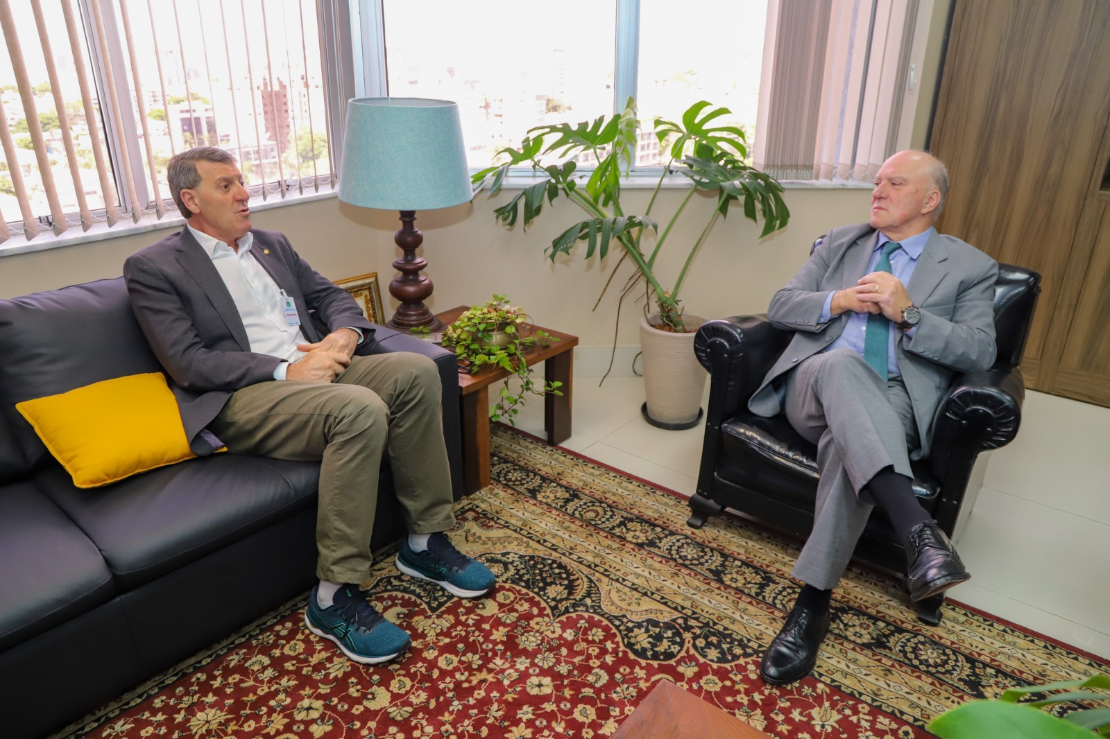 Na foto, duas pessoas estão sentadas em uma sala. À esquerda, um homem veste um blazer cinza escuro, camisa branca e calça bege, além de tênis azul escuro. Ele está sentado em um sofá preto com uma almofada amarela ao lado. À direita, outro homem usa um terno cinza, gravata verde e sapatos pretos. Ele está sentado em uma poltrona de couro preta e mantém as mãos juntas sobre o colo.