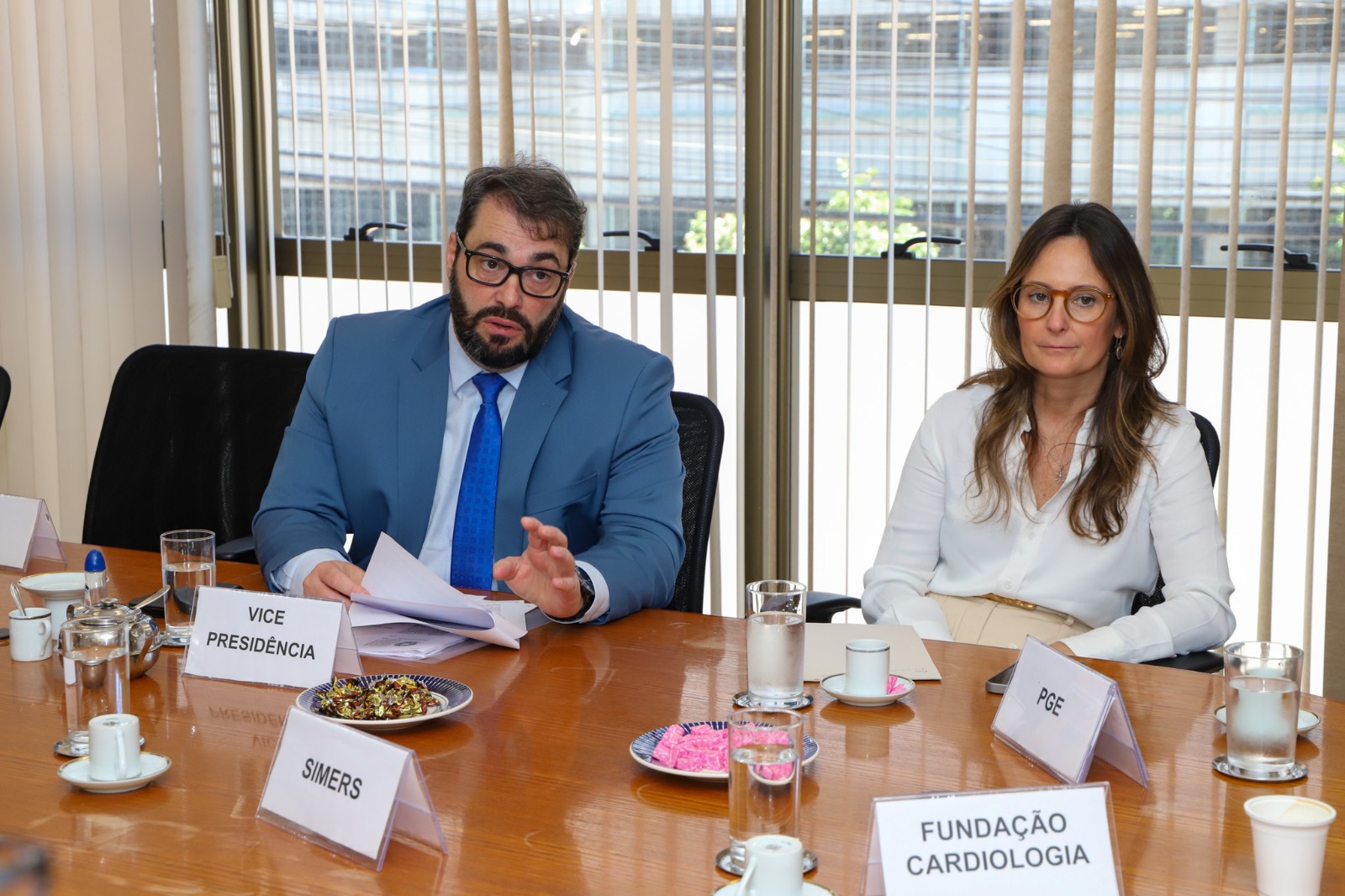 A imagem mostra um homem de terno azul, camisa branca e gravata azul ao lado de uma mulher que veste uma blusa branca. Ambos estão sentados ao redor de uma mesa. 