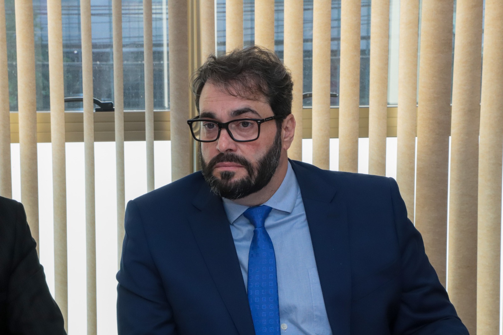 A imagem mostra um homem de barba, usando óculos e com terno azul e gravata azul. E camisa clara.