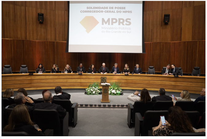 A imagem retrata a solenidade de posse do Corregedor-Geral do Ministério Público do Rio Grande do Sul (MPRS). No palco, autoridades estão sentadas em uma mesa em formato semicircular. Um painel de fundo exibe o logo do MPRS e o título do evento. A plateia assiste ao ato.