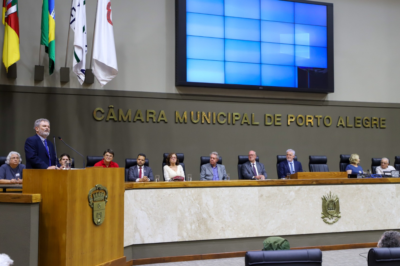 A imagem mostra uma sessão na Câmara Municipal de Porto Alegre. Há várias autoridades sentadas em uma mesa longa, com Castrinho de pé falando em um púlpito à esquerda.
