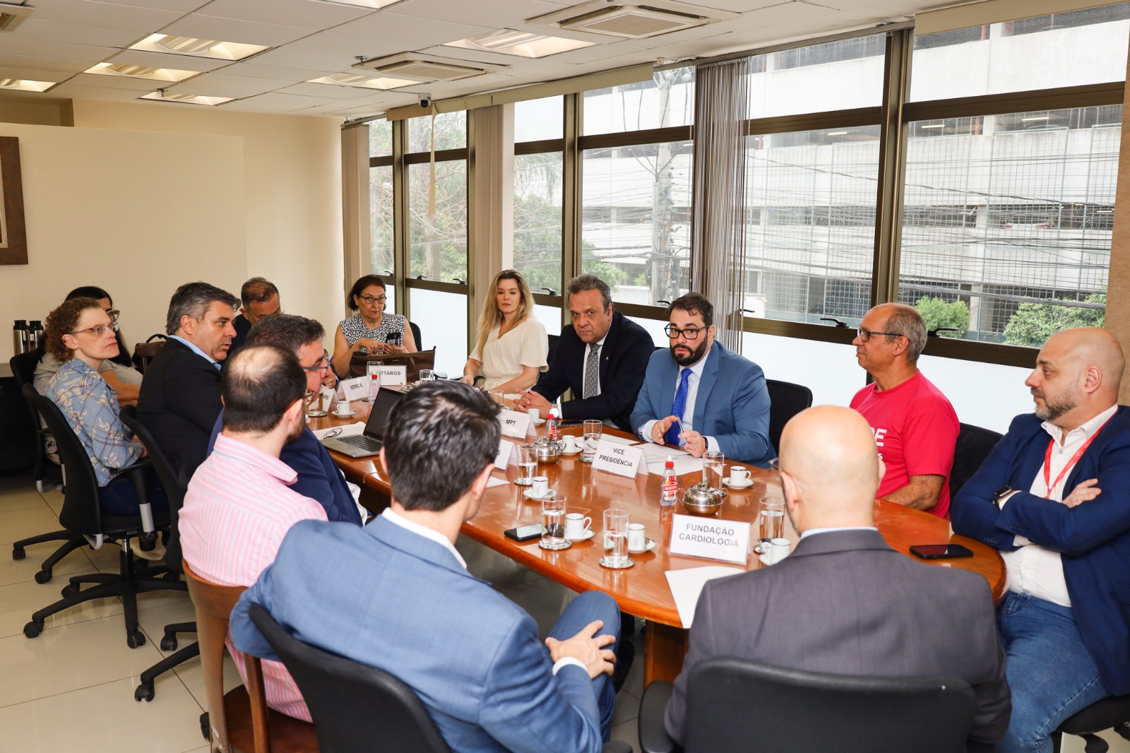 A imagem mostra uma mediação com várias pessoas ao redor de uma mesa de conferência, com documentos e laptops. As placas de identificação incluem "VICE PRESIDÊNCIA," "FUNDAÇÃO CARDIOLOGIA," e "ABP."