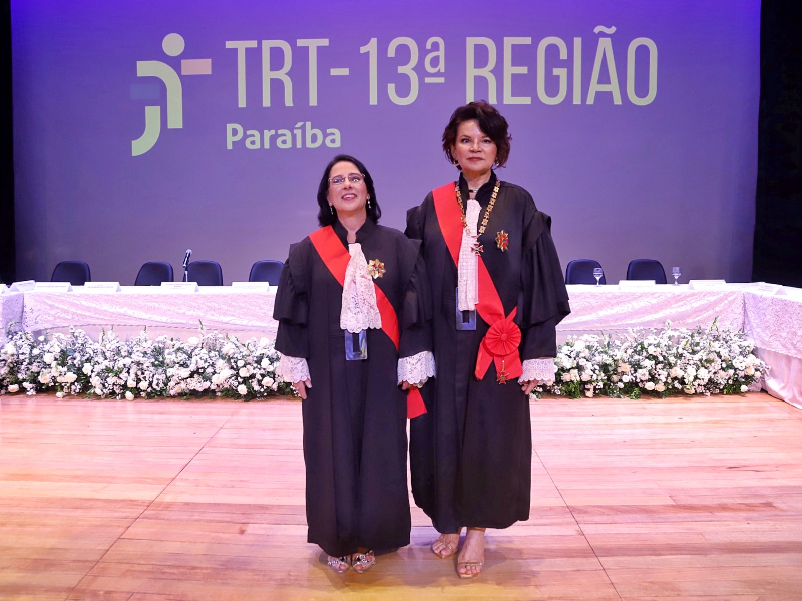A imagem mostra as empossadas com trajes formais, incluindo togas pretas com detalhes em renda branca e faixas vermelhas cruzadas no peito. Uma delas também usa uma corrente dourada ao redor do pescoço. Elas estão em pé sobre um palco de madeira, em frente a uma mesa comprida coberta com uma toalha branca e decorada com arranjos de flores brancas. Ao fundo, há um painel com o texto "TRT - 13ª REGIÃO Paraíba".