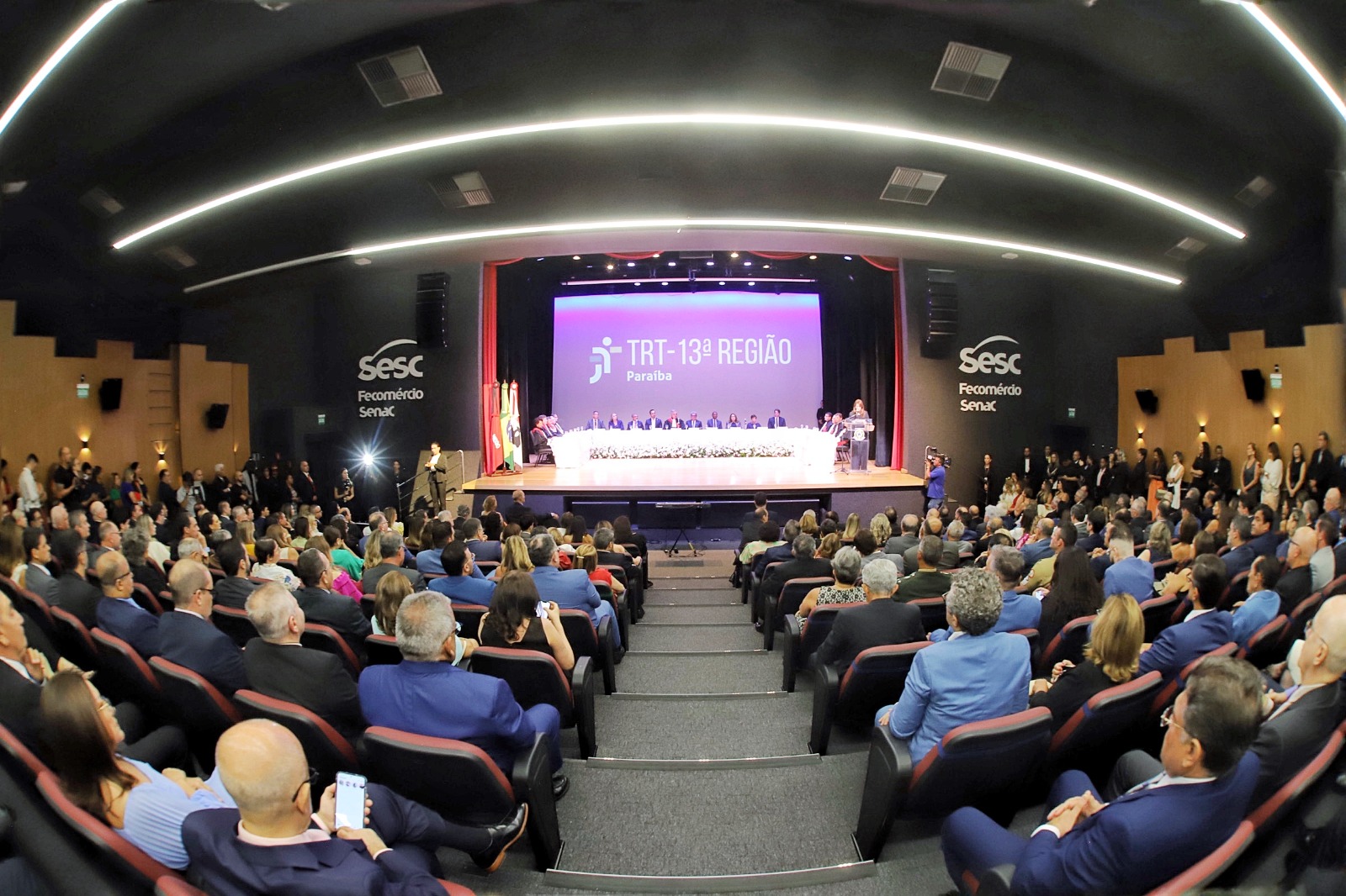 A imagem mostra um auditório cheio de pessoas assistindo a um evento formal. No palco, há uma mesa longa com várias pessoas sentadas atrás dela, e uma pessoa está em pé, aparentemente fazendo um discurso. No fundo do palco, há um grande painel roxo com o texto "TRT - 13ª REGIÃO Paraíba". O auditório está bem iluminado e as pessoas estão sentadas em cadeiras vermelhas e pretas, prestando atenção ao que está acontecendo no palco.