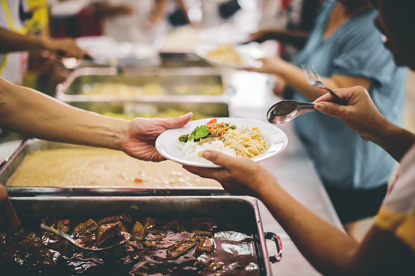 A imagem mostra uma cena em que pessoas estão servindo comida em um ambiente de refeitório.