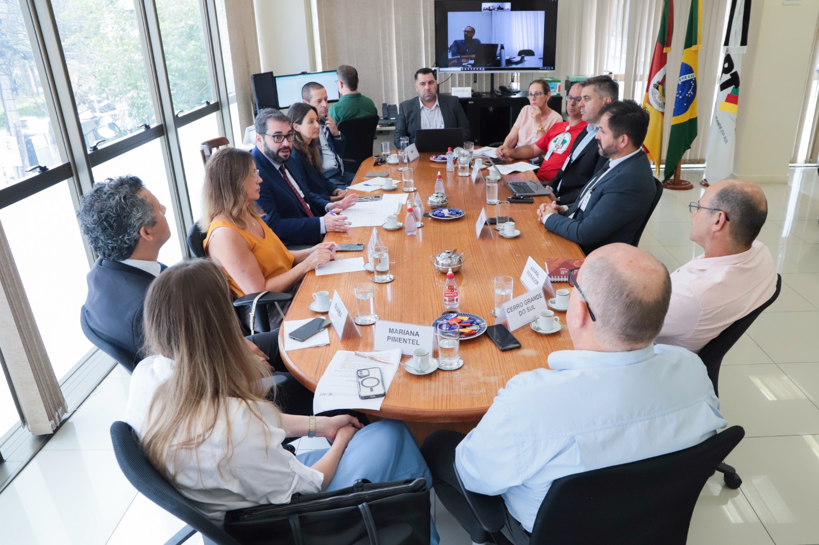 Foto dos participantes na mesa de negociação. Vice-presidente aparece falando, no lado esquerdo. Ao fundo há uma tela de videoconferência ligada e as bandeiras do RS, do Brasil e da Justiça do Trabalho