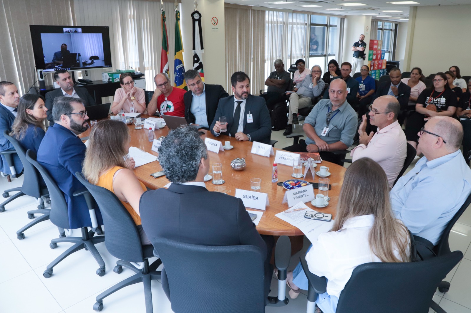 Foto da mediação com a mesa de negociação em primeiro plano e um público ao fundo assistindo.