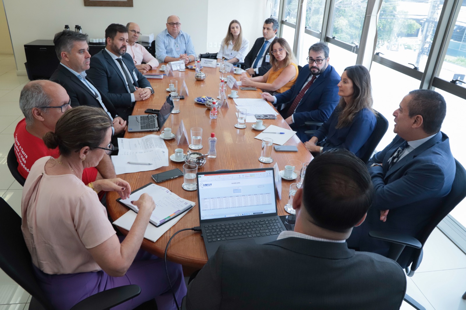 Foto dos participantes na mesa de negociação pelo ângulo inverso. Ao fundo aparece a parede e do lado direto a janela. Há treze pessoas na imagem.