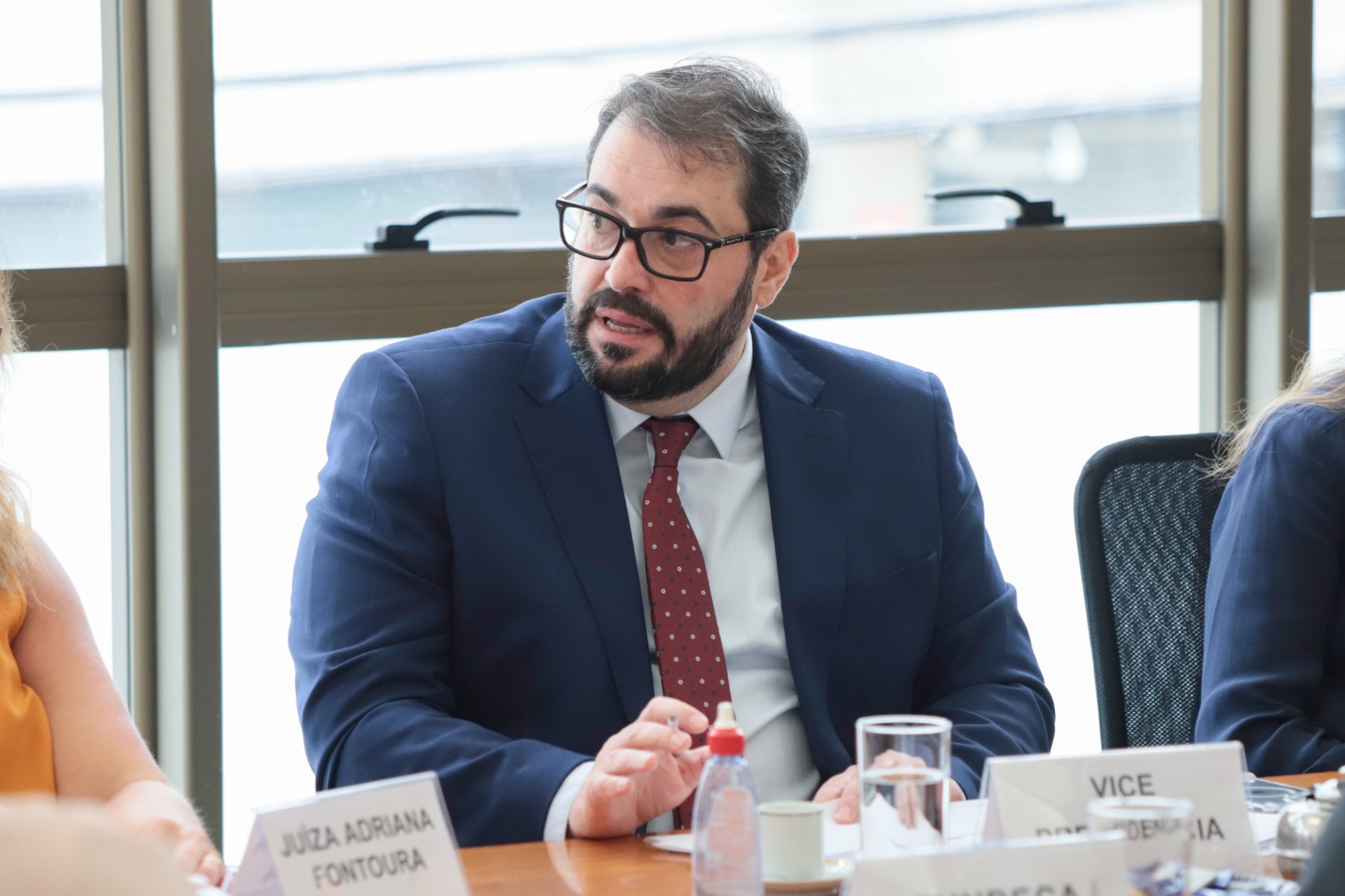 Foto do vice-presidente Alexandre Correa da Cruz, Ele veste um terno azul marinho, fala, e está com o rosto voltado para a direita. Usa barba e óculos de armação escura. É branco e de cabelo escuro.