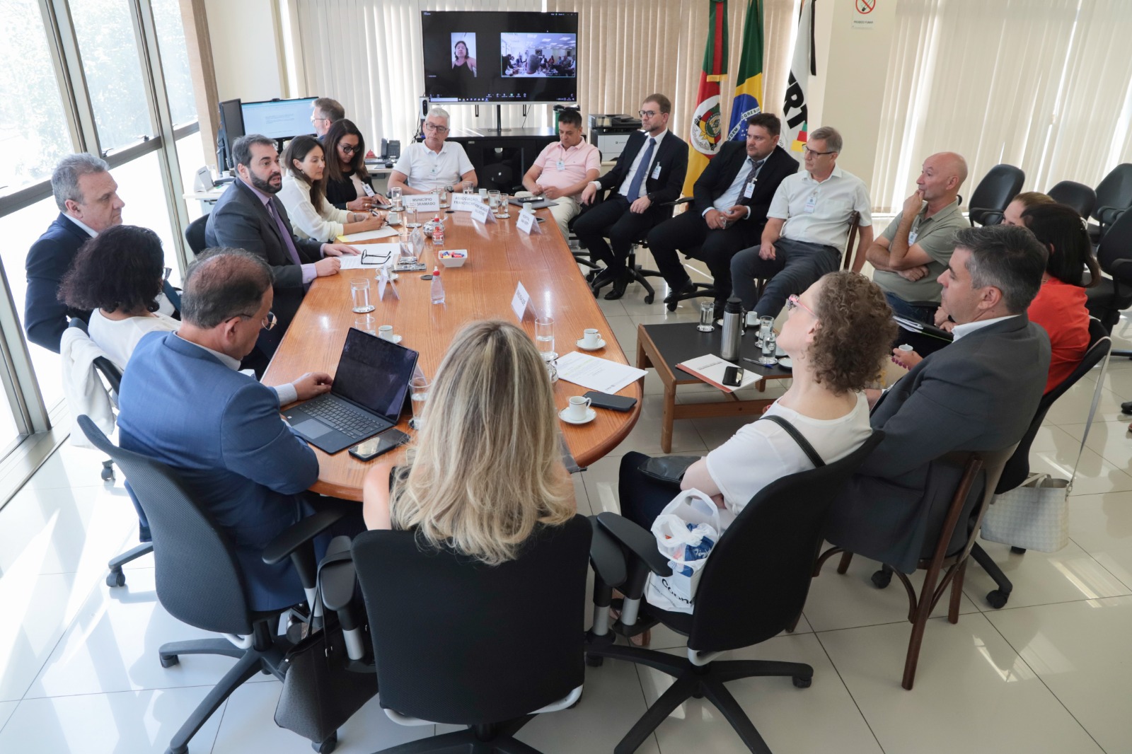 Foto das pessoas na mesa de negociação. Ao fundo, aparece uma tela de videoconferência e as bandeiras do RS, do Brasil e da JT. 18 pessoas aparecem na imagem. As cadeiras estão disposta em circulo, algumas um pouco mais afastadas da mesa para todos conseguirem participar.