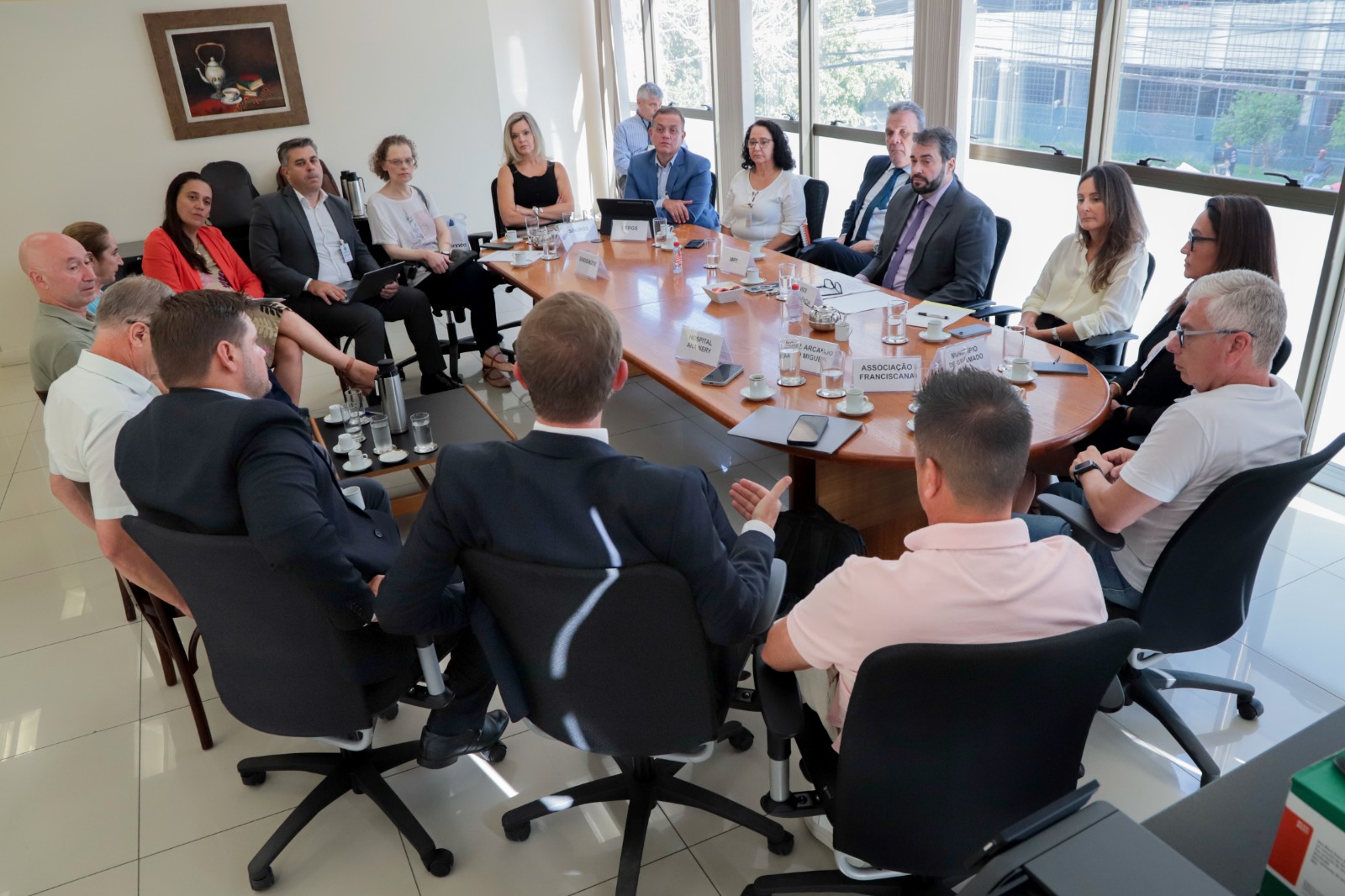 Foto das pessoas na mesa de negociação pelo ângulo inverso, com um quadro na parede banca ao fundo.