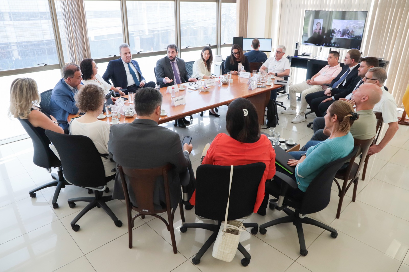 Foto das pessoas na mesa de negociação por outro ângulo, com a janela da sala ao fundo.