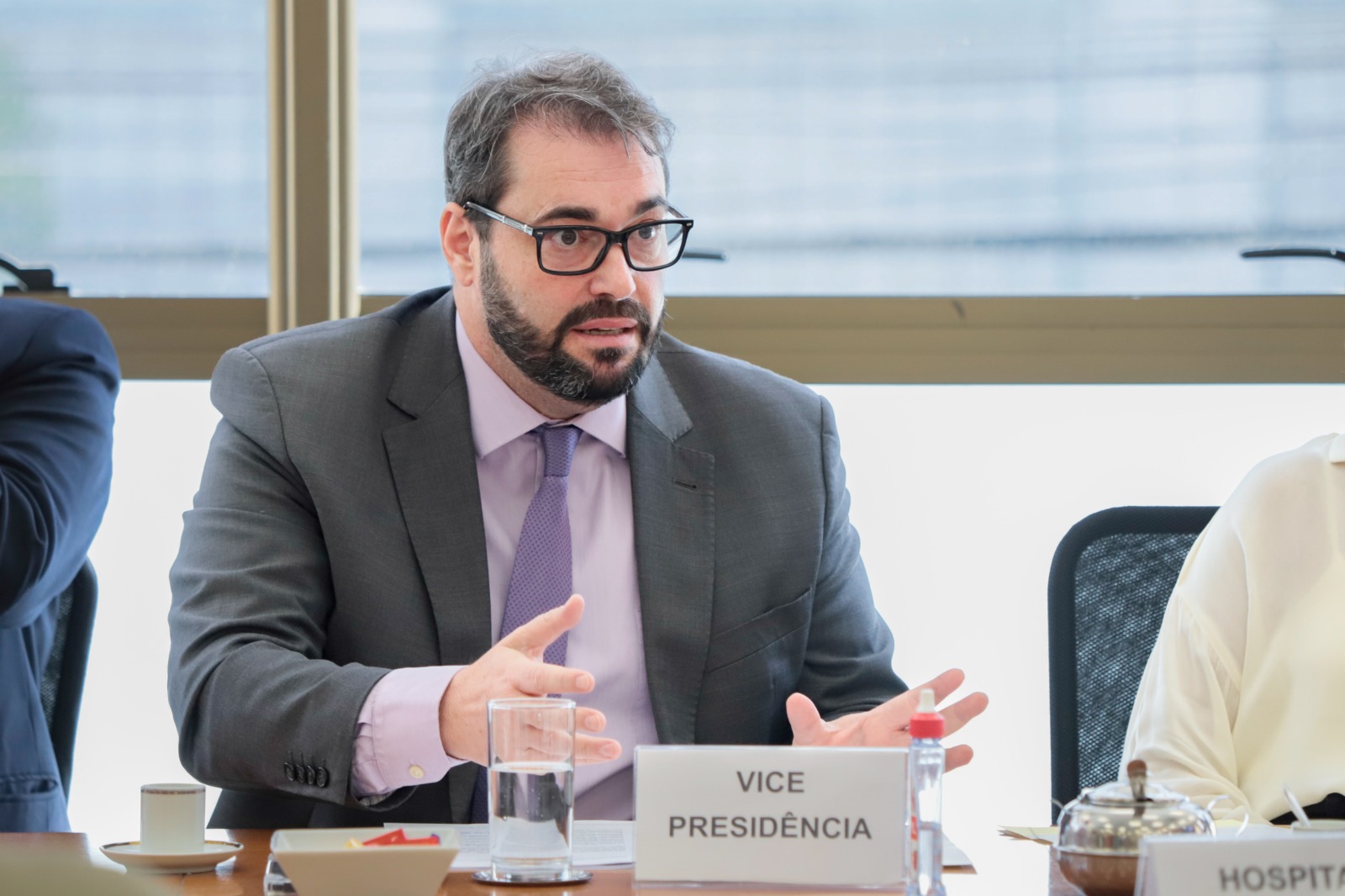 Vice-Presidente Alexandre Corrêa da Cruz fala e gesticula. Ele é branco, usa cabelo e barba pretos e óculos de armação preta. À sua frente na mesa aparece uma placa onde está escrito "Vice-Presidência".