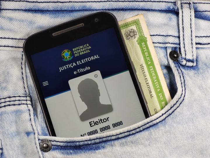 Celular com o aplicativo e-título aberto e um título eleitoral em papel dentro de um bolso de calça jeans.Texto: Decisão.