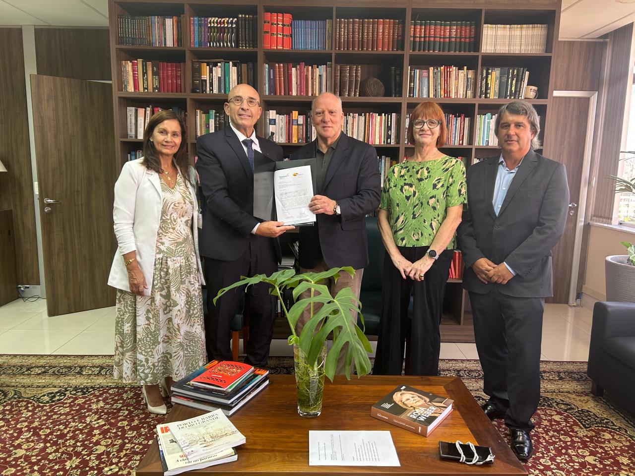 Os 5 participantes da reunião posam para a foto no gabinete do presidente. O espaço conta com estante de livros ao fundo. Na frente, uma mesa de madeira com livros e uma planta.