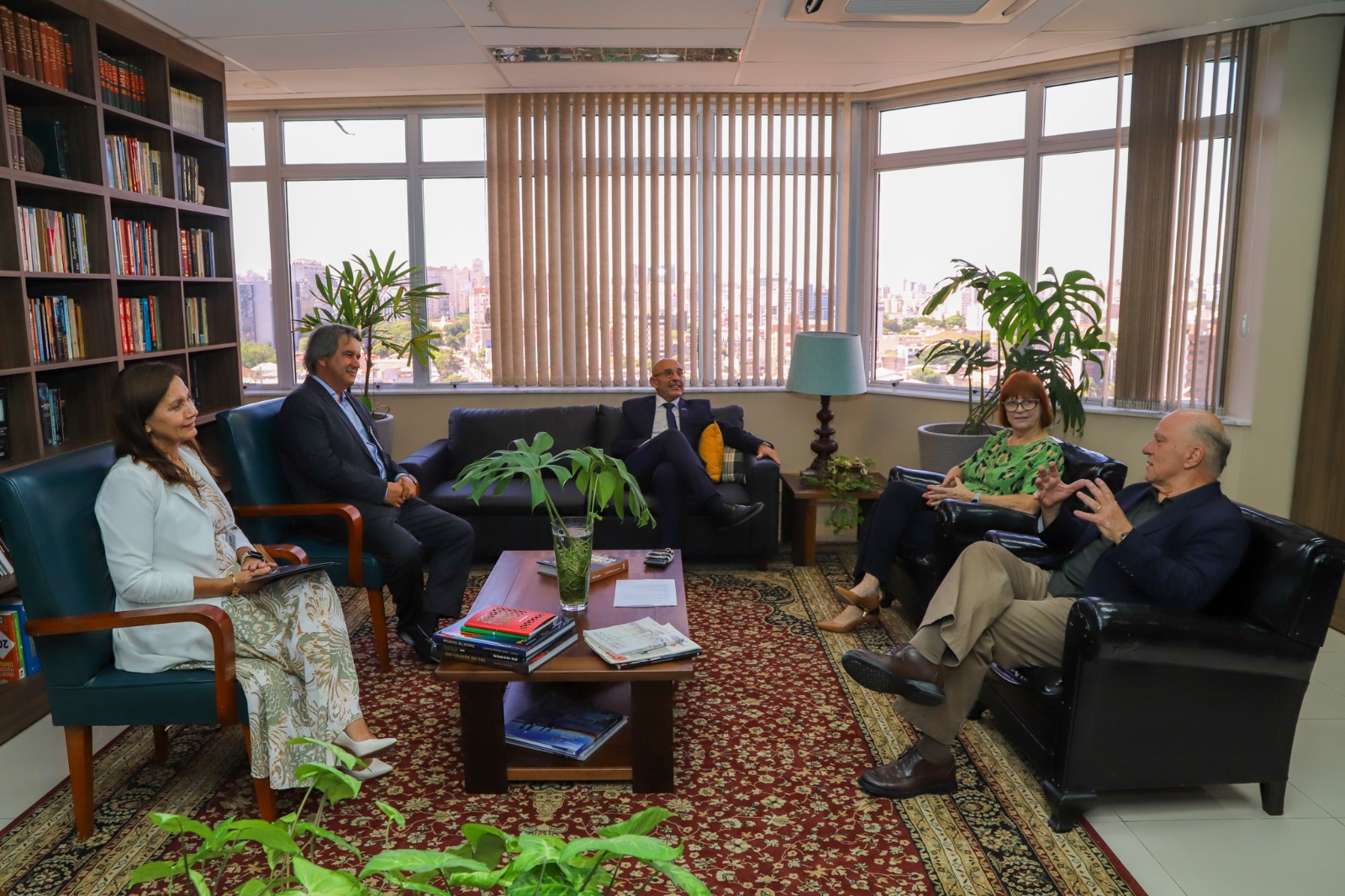 Os cinco participantes da reunião estão sentados no gabinete do presidente. O ambiente é iluminado por janelas amplas. Há poltronas, uma mesa de centro com livros e plantas decorativas. Ao fundo, é possível ver a vista da cidade.