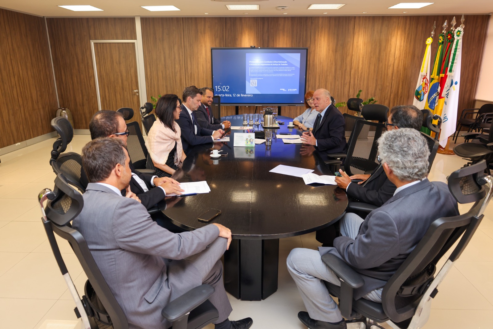 Participantes da reunião estão sentados ao redor de uma mesa, discutindo assuntos. Há papéis, copos de café e uma tela ao fundo.