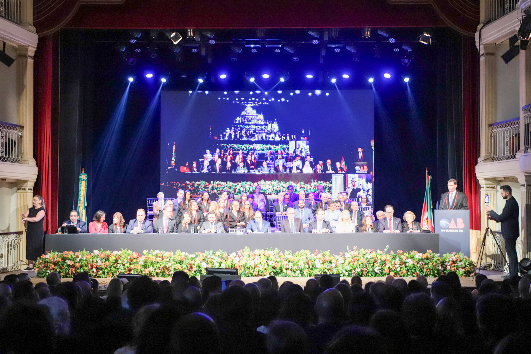 Foto em ângulo aberto da cerimônia, mostrando o público em primeiro plano, e o palco ao fundo. Foto tirada no momento em que Leonardo Lamachoa discursava no púlpito.